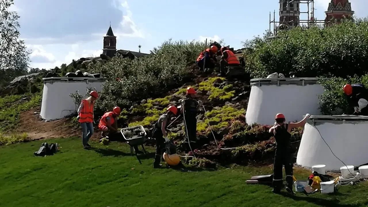 «Зарядье», Москва, 2017 · посадочные работы на склоне Рабочие в касках и жилетах сажают растения на склоне, рядом бетонные кольца и тачка